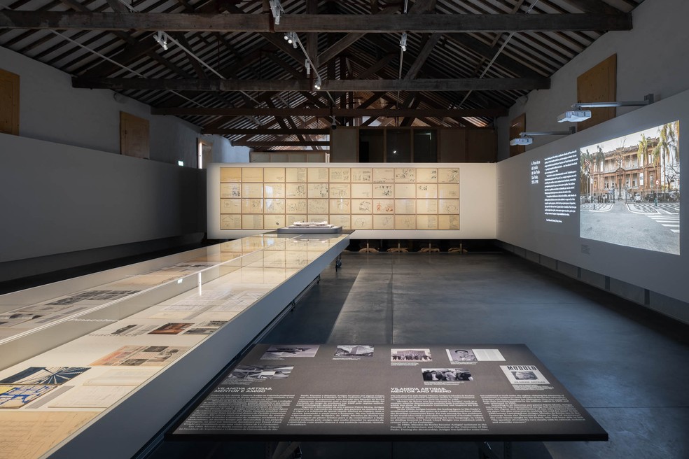 Os materiais de arquivo são apresentados numa mesa central, enquanto as paredes possuem grandes projeções que mostram aos visitantes a vida cotidiana dos edifícios — Foto: Ivo Tavares Studio/Casa da Arquitectura