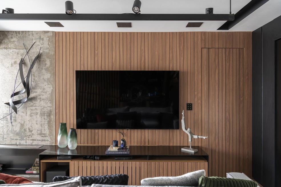 Para emoldurar a TV, a arquiteta desenhou um painel de madeira que dialoga com o armário sob medida - na lateral, a parede em concreto aparente reforça a inspiração industrial do projeto — Foto: Cris Farhat