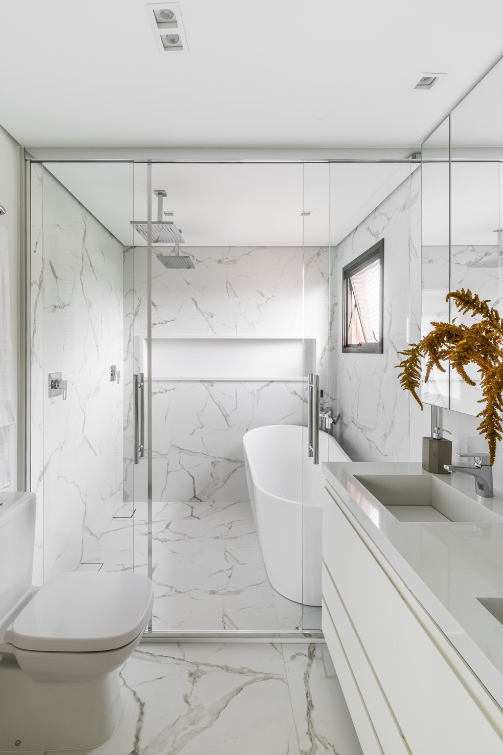Projetado pela arquiteta Vivi Cirello, esse banheiro é todo revestido por pedras marmorizadas e possui uma bancada e nicho da mesma pedra em branco — Foto: Divulgação