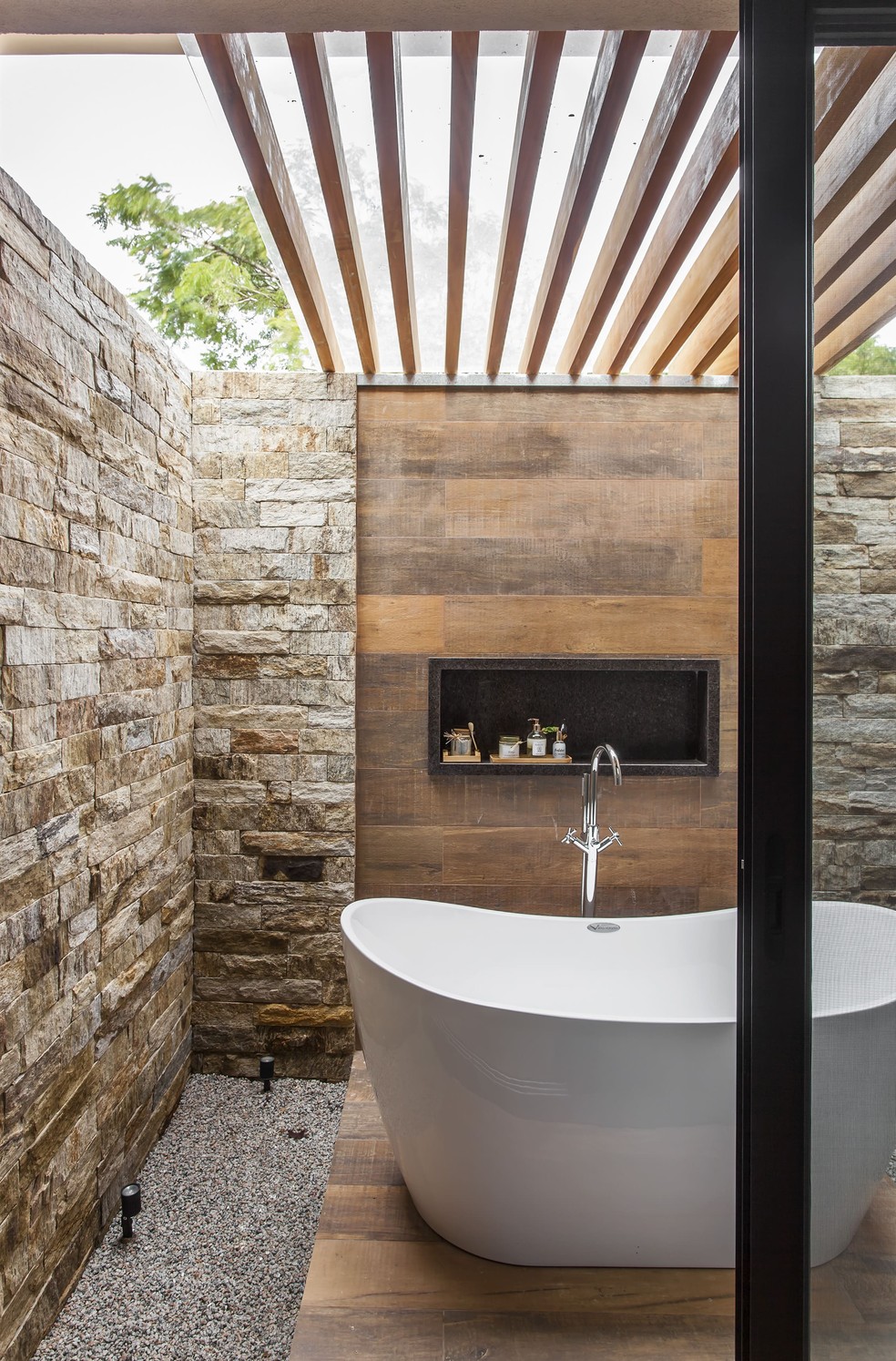 Banheiro ao ar livre, com paredes de pedra madeira, nicho de pedra marrom e banheira branca, projetado pelo escritório Korman Arquitetos — Foto: Eduardo Pozella