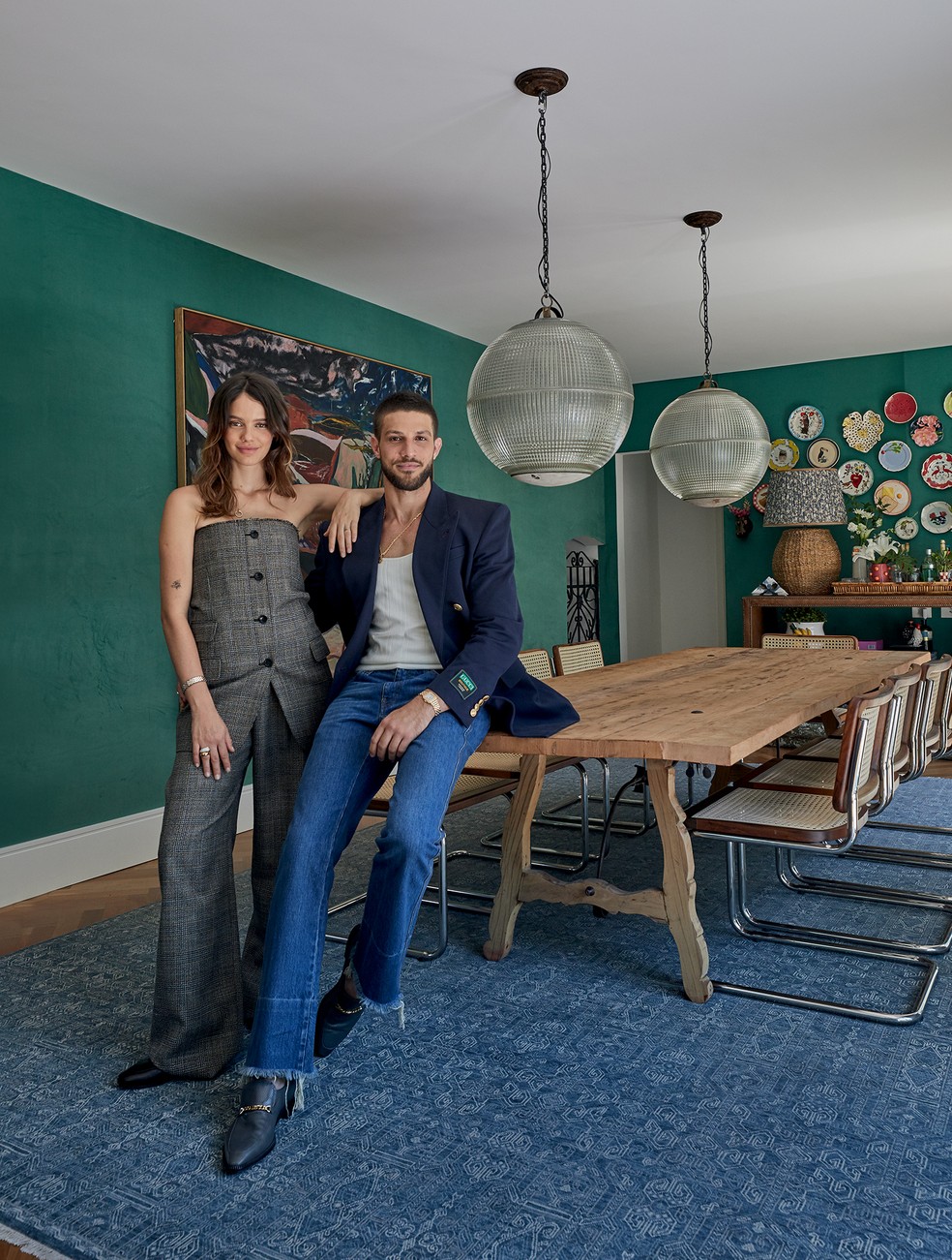 Na sala de jantar, Laura veste Gucci e joias Cartier e posa ao lado de Chay, com look Gucci, colar Alan Crocetti, relógio e jóias Cartier: o ambiente foi decorado com pendentes do metrô de Paris dos anos 1940 e mesa de jantar espanhola do séc. 19, tudo na Galeria Juliana Benfatti, e cadeiras vintage Cesca, de Marcel Breuer, no antiquário Bossa Nossa Casa, sobre tapete da By Kamy, enquanto as paredes, com veludo verde da coleção Majorelle, da Entreposto, ostentam tela de Fernanda Galvão e coleção de pratos garimpados pela moradora (boa parte da Anthropologie) – ao fundo, abajur da Entreposto — Foto: Deco Cury