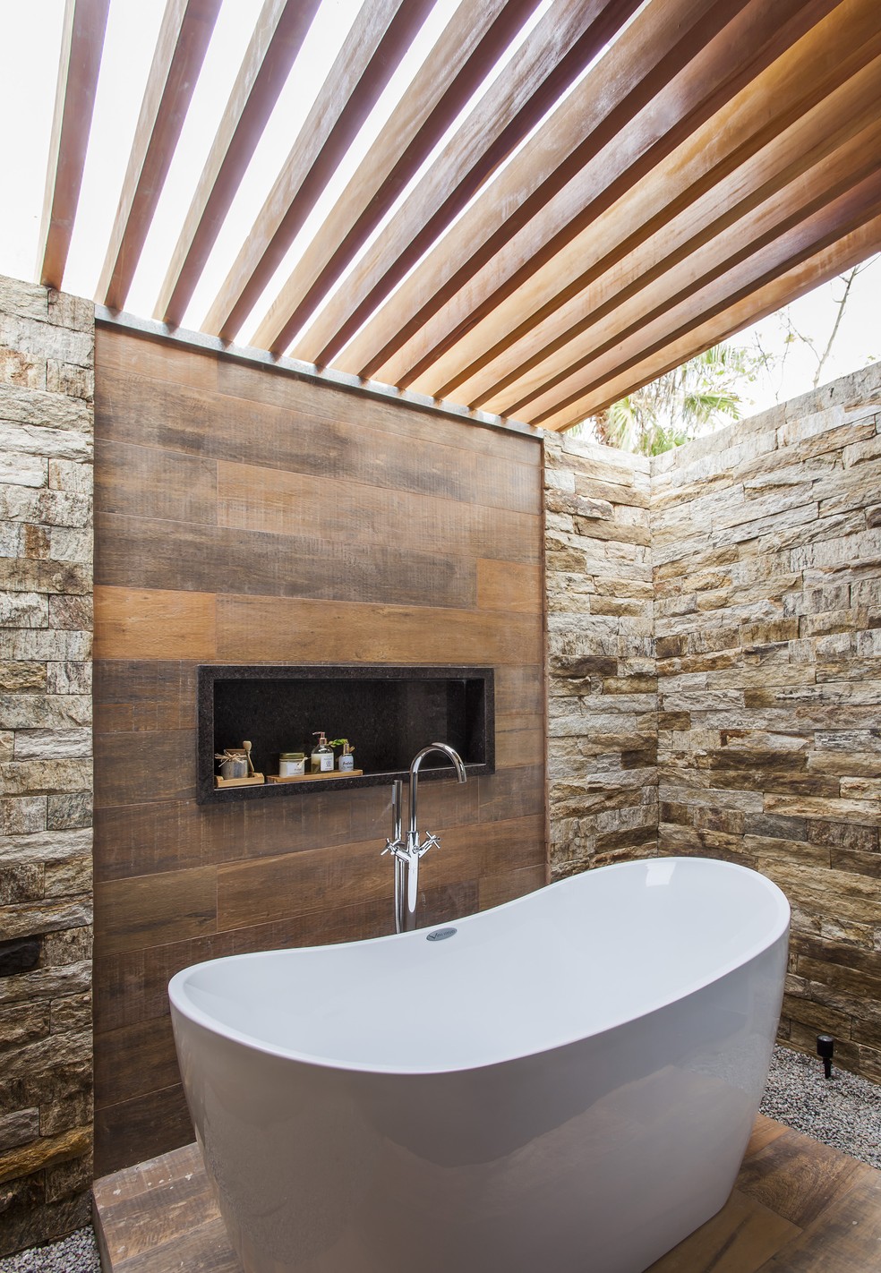 Com o projeto do escritório Korman Arquitetos, esse banheiro ao ar livre possui um nicho embutido em pedra marrom e com o teto fechado  na parte da banheira com pergolado de madeira e vidro — Foto: Eduardo Pozella