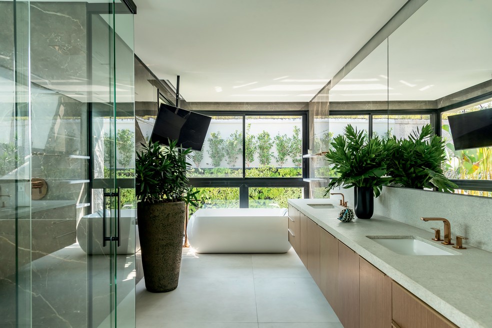 O banheiro da suíte máster ganhou vários itens visando conforto e bem-estar, como a TV, a banheira e o box de vidro executado pela Vidros Quitaúna – além da vista para o jardim — Foto: Wesley Diego Emes