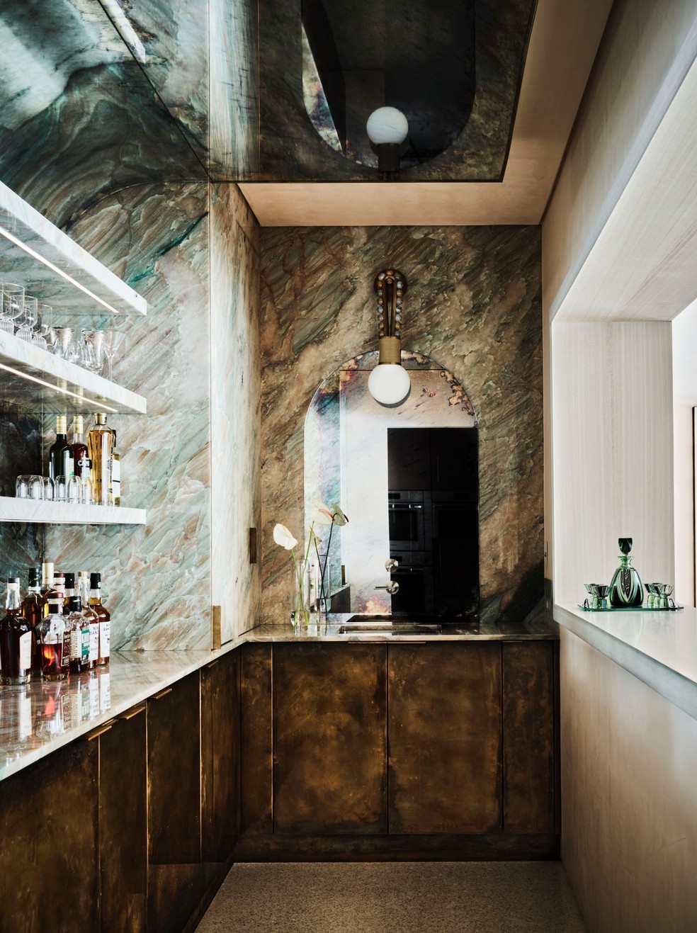 A arandela da Apparatus (ao centro) dá o tom da área do bar, com espelho, revestimento de quartzito e armários de latão patinado — Foto: Douglas Friedman