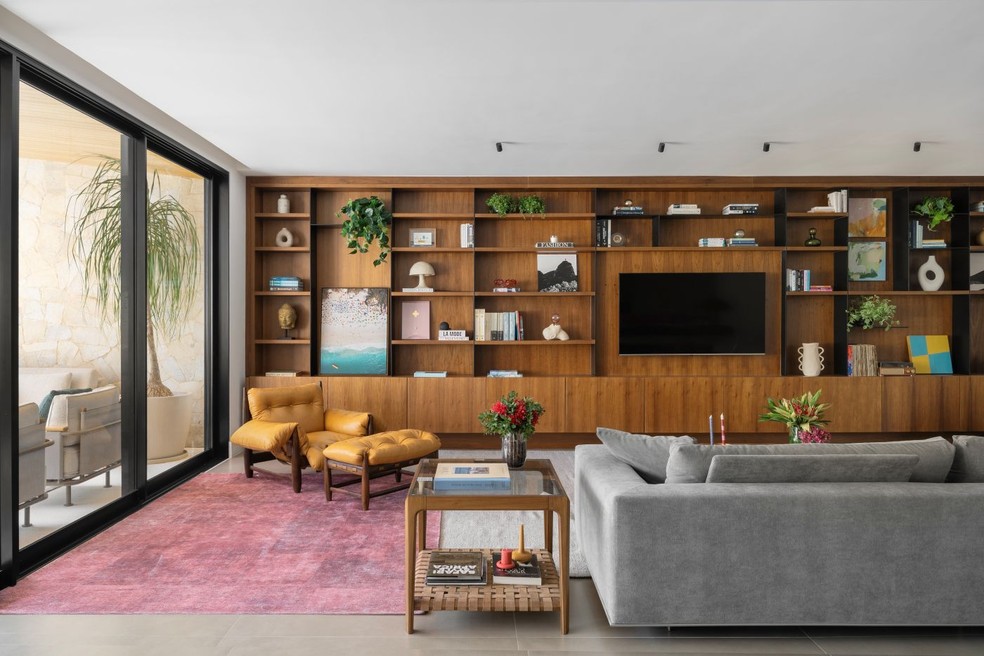 Projetada pelo Flavia Malvaccini Arquitetura, esta sala de TV ganha aconchego por meio da estante de madeira, decorada com obras de arte, abajures e pilhas de livros — Foto: Estúdio NY18/Divulgação