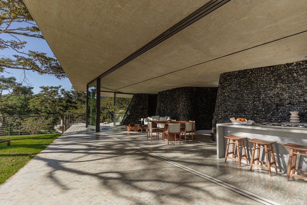 Mais um ângulo da área social toda emoldurada pela parede de pedras portuguesas preta -- em primeiro plano, as banquetas Bar Nine, de Sergio Rodrigues, e, mais ao fundo, a mesa de jantar ganha a companhia das cadeiras Cantú, de Sergio Rodrigues, tudo na São Romão  — Foto: Luisa Lage