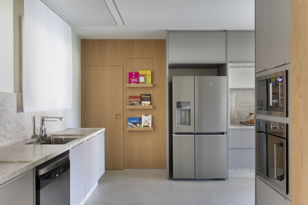 Com base em tons de cinza, madeira foi utilizada em diversas áreas da cozinha e copa - como ao lado da geladeira, com estantes para livros — Foto: Juliano Colodeti/MCA Estúdio