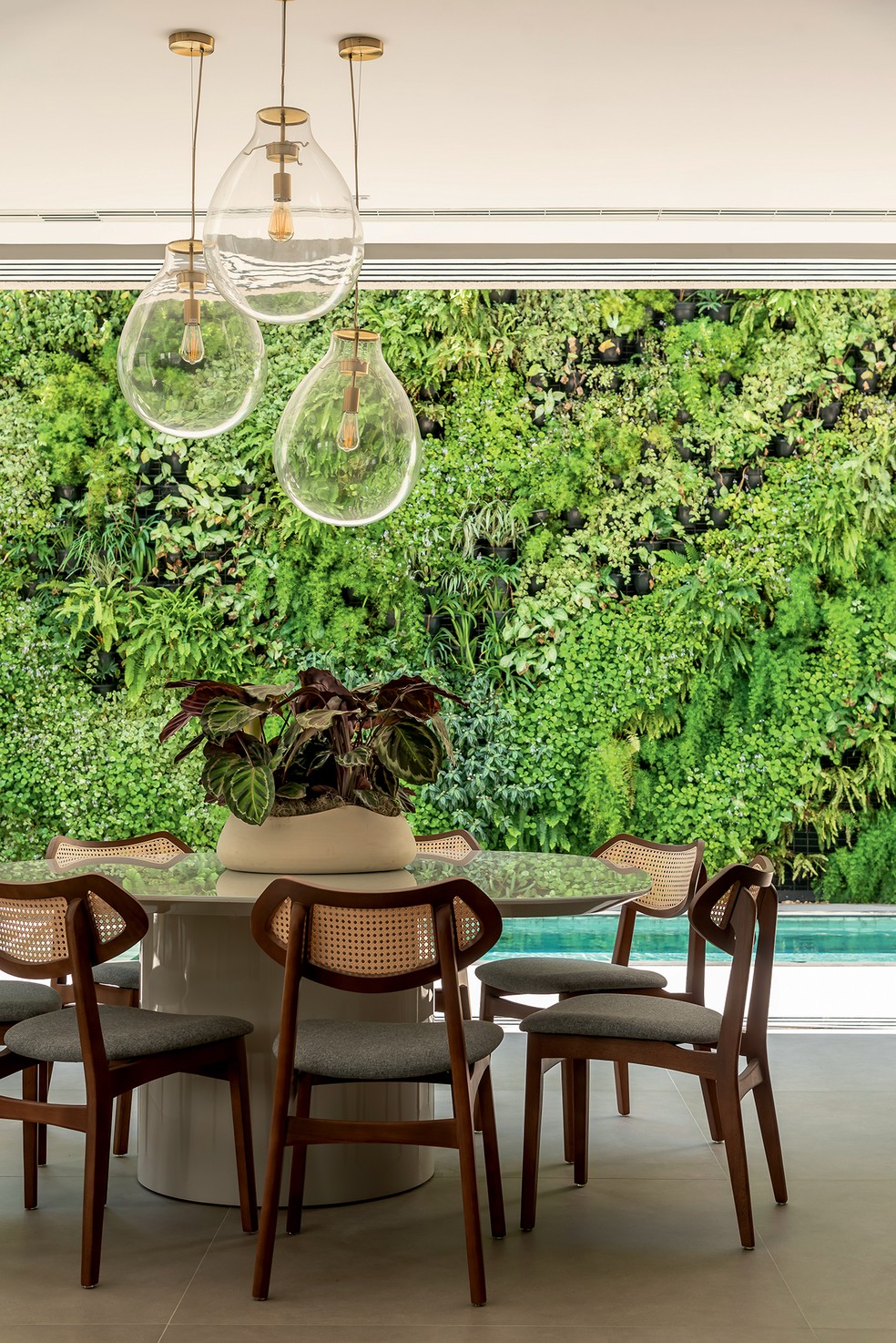 Com vista para o exuberante jardim vertical e a piscina, o espaço gourmet foi ambientado com mesa e cadeiras feitas de palhinha, madeira e tecido, e pendentes da Perfeita Luz, que combinam vidro e metal — Foto: Wesley Diego Emes